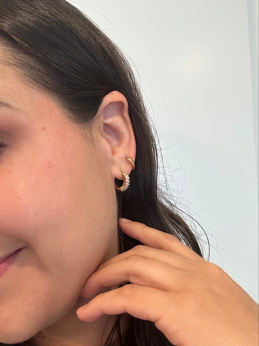 A close-up side view of a woman's ear wearing two small gold hoop earrings, with her hand near her neck against a plain light background.