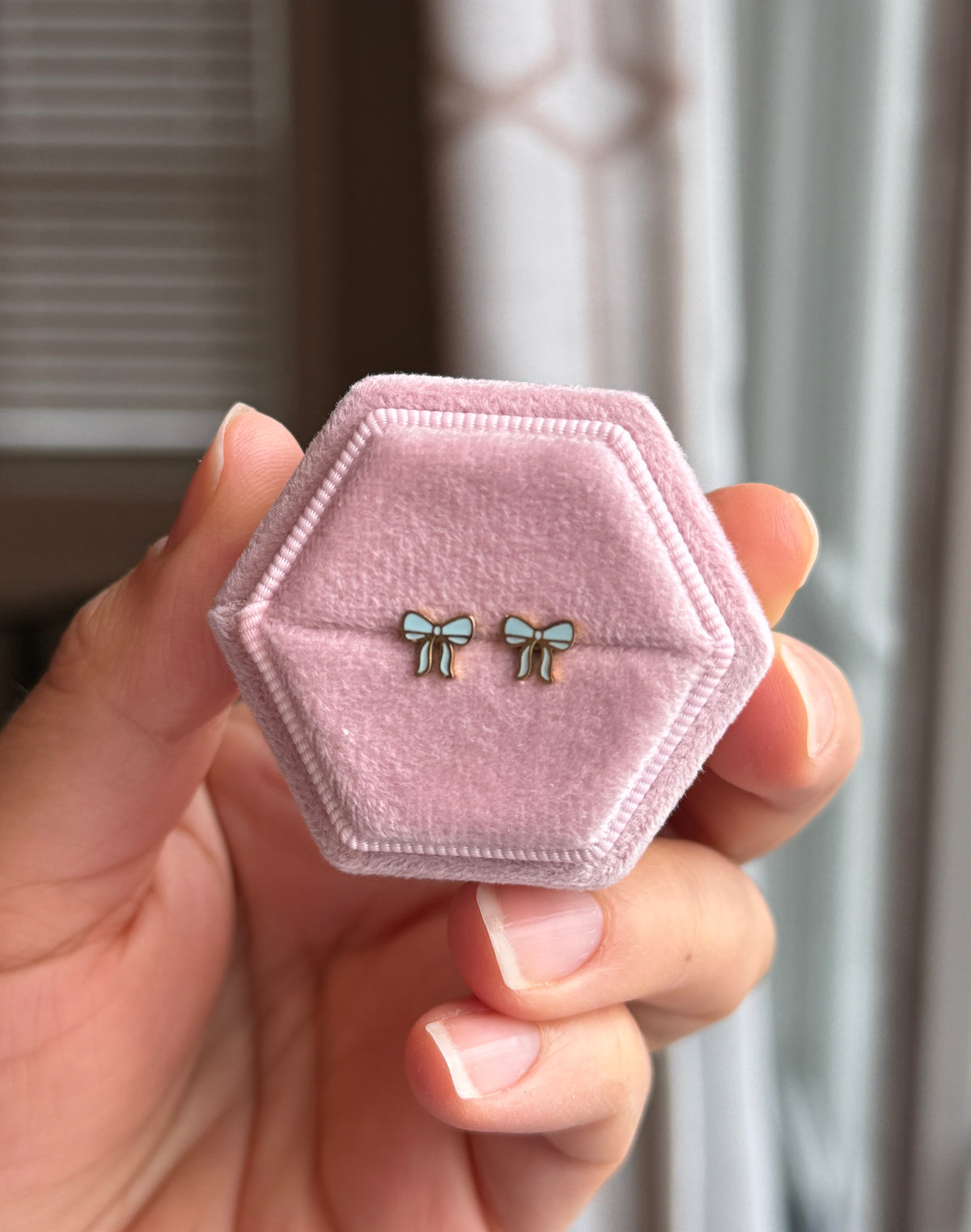 Baby blue and gold bow studs shown on a light pink velvet jewelry box held by a hand