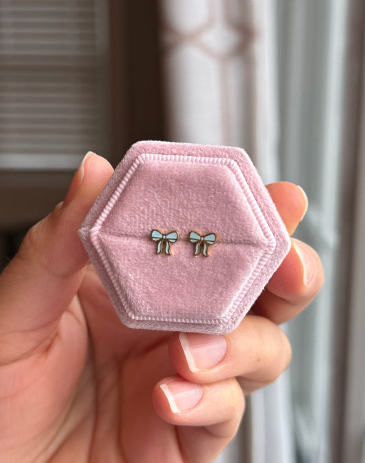 Baby blue and gold bow studs shown on a light pink velvet jewelry box held by a hand