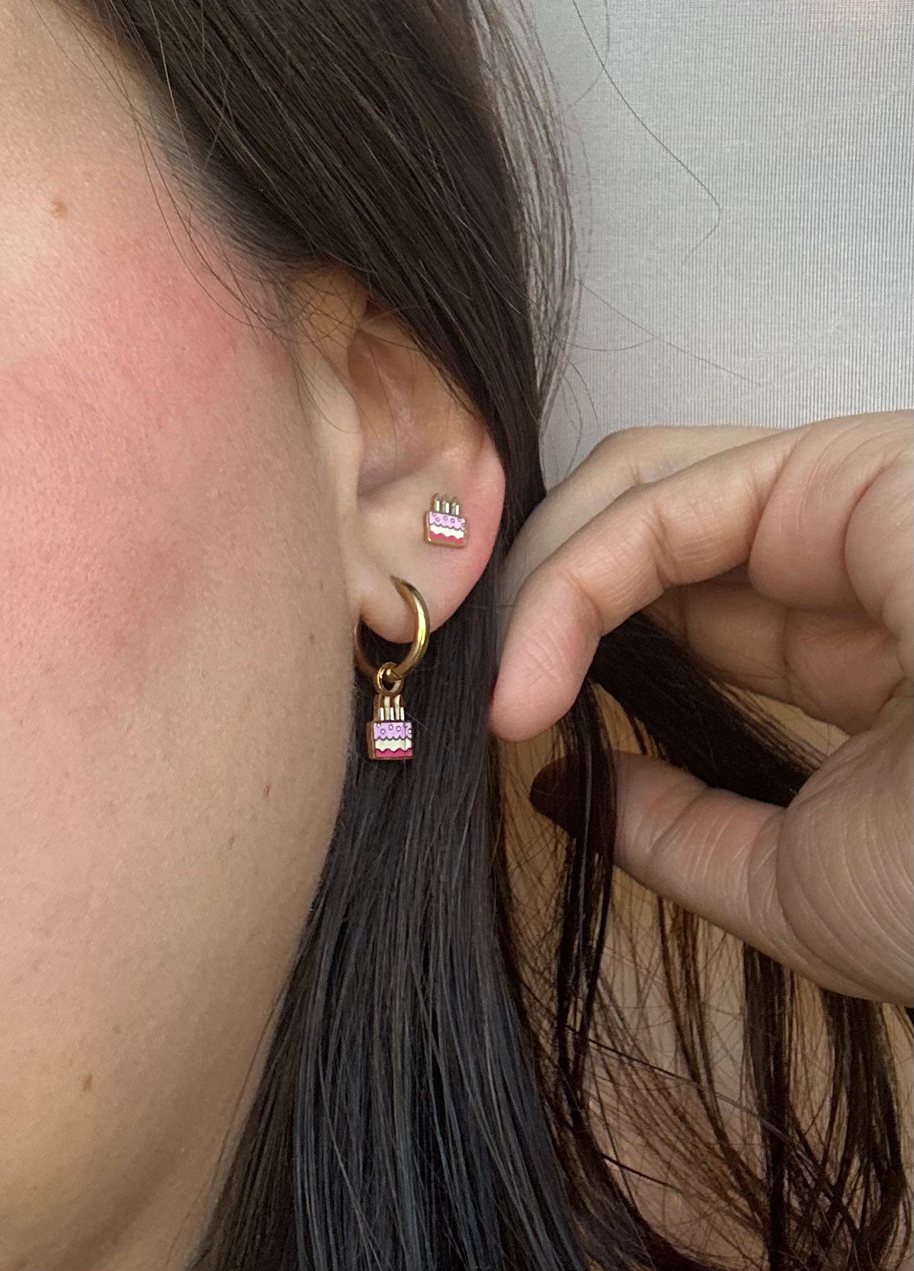 Birthday cake studs stacked with birthday cake charm hoops on an ear to show size
