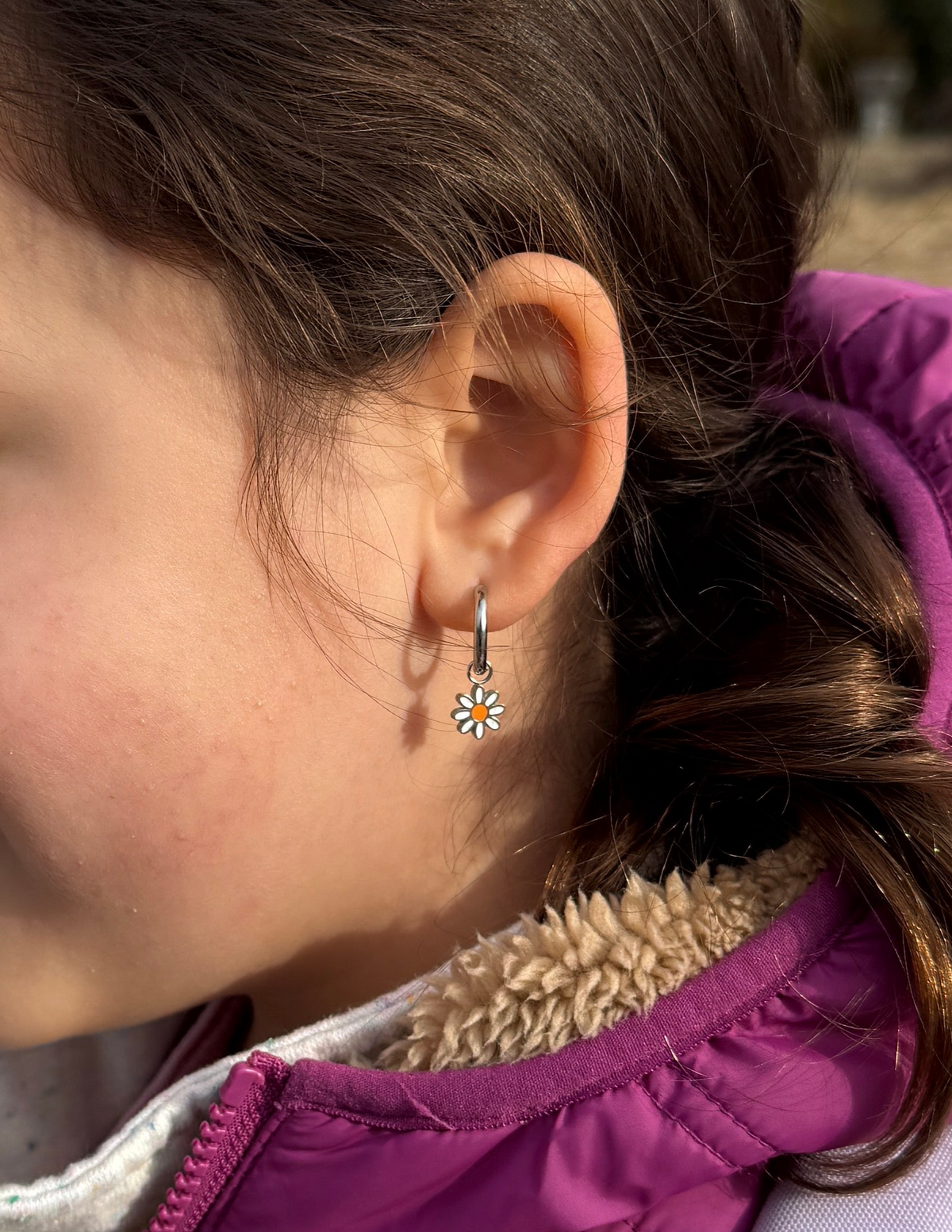 A close-up side profile of a young girl wearing a small silver daisy drop earring, wearing a purple puffer jacket with a beige fleece lining.