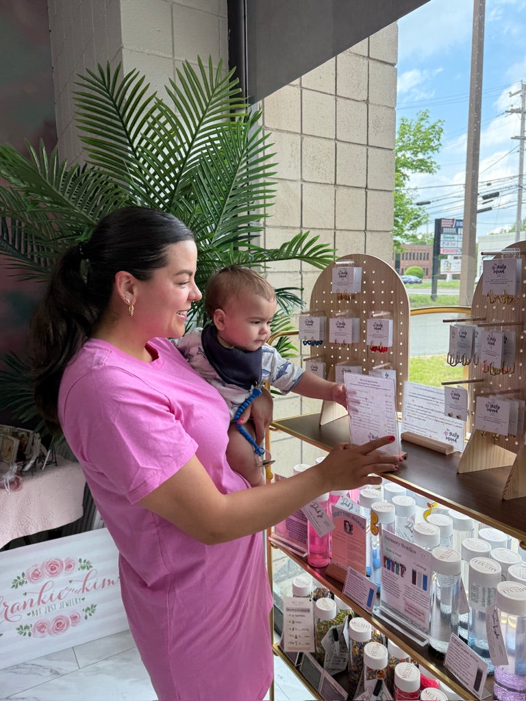 A woman in a pink shirt dress holds a baby while looking at earrings displayed on shelves inside a boutique.